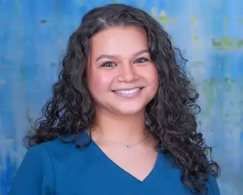 Smiling female dental staff member with long dark curly hair wearing blue scrubs.