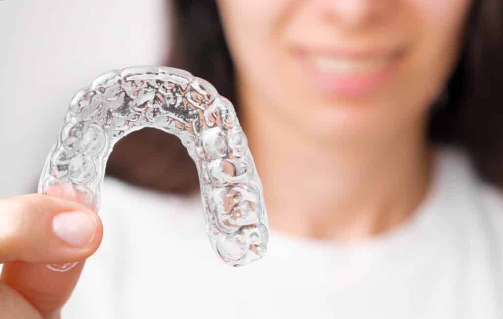 A person holds a clear dental aligner in focus, smiling softly in the background. The aligner is transparent and curved, reflecting light.